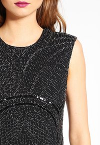 Svart ärmlös topp med intrikata pärldetaljer och texturmönster, med en rund halsringning och slät tyg.
