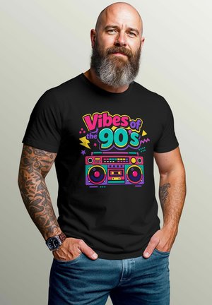 Bärtiger Mann mit Tattoos, der ein schwarzes T-Shirt mit buntem "Vibes of the 90's"-Schriftzug und einer Retro-Boombox-Grafik trägt, Hände in den Taschen der blauen Jeans.