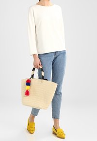 Woven straw tote bag with black and white striped handles, adorned with colorful tassels in red, yellow, and blue. Model is dressed in casual attire.