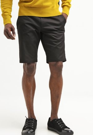 Homme portant un short noir jusqu'aux genoux, un pull jaune et des baskets noires basses à lacets, debout avec une main dans la poche.