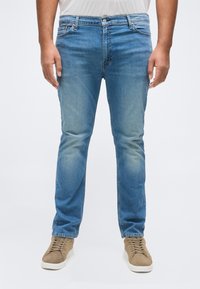 Homme portant un jean bleu slim et des baskets marron clair, debout avec les bras détendus le long du corps sur fond blanc.