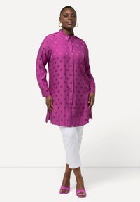 Chemise à manches longues, à boutons, de couleur violet vif avec de grands pois. Le tissu est lisse, avec des fentes sur les côtés. Portée sur un pantalon blanc court.