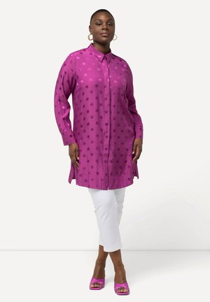 Chemise à manches longues, à boutons, de couleur violet vif avec de grands pois. Le tissu est lisse, avec des fentes sur les côtés. Portée sur un pantalon blanc court.