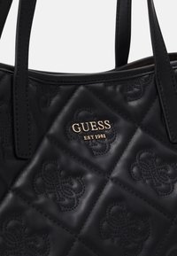 Sac à main noir matelassé avec motifs embossés, poignées noires et logo Guess doré avec "EST 1981" à l'avant.