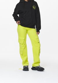 Černá mikina s kapucí s kruhovou grafikou, doplněná vysoce viditelnými limetkovými cargo kalhotami s několika kapsami. Celý outfit dotvářejí tenisky.