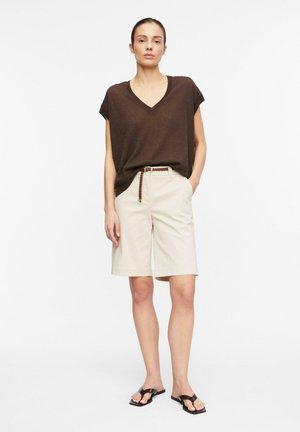 Kobieta stoi, ubrana w brązowy krótki top z dekoltem w serek, beżowe szorty do kolan z plecionym paskiem i czarne sandały z odkrytymi palcami.