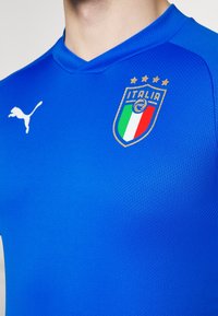 Niebieski sportowy strój wykonany z teksturowanej tkaniny. Posiada dekolt w serek, logo Puma oraz herb narodowej drużyny Włoch z złotymi gwiazdami.