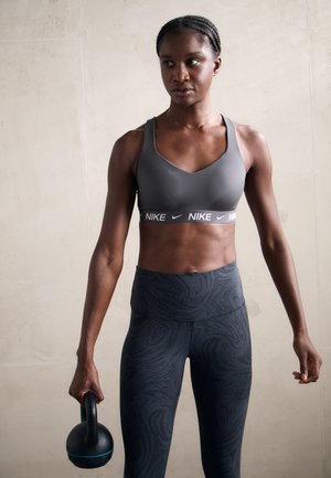 Grå sportbh med hvit Nike-logo og svarte høytalende leggings med svirle mønster; holder en svart kettlebell med blå detalj.