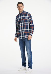 Camicia a quadri con tonalità di blu navy, bianco e arancione, colletto button-down e due tasche frontali, abbinata a jeans blu e sneakers bianche.