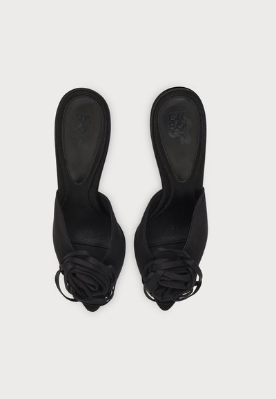 Paire de chaussures à talons hauts noires à bout ouvert avec détail floral sur le devant et fines brides à la cheville, vues de dessus sur fond blanc.