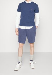 Tmavě modré tričko s krátkým rukávem a kapsou na prsou, vrstvené přes bílé košili s knoflíky. Světle modré šortky a bílé tenisky se šedými ponožkami.