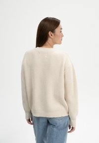 Beiger gestrickter Pullover mit lockerer Passform, gerippter Textur und Rundhalsausschnitt, kombiniert mit hellblauen Jeans. Sichtbare Nahtdetails am Saum und an den Bündchen.