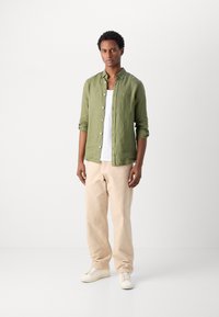 Cămașă ușoară din in verde, top alb pe dedesubt, pantaloni din bumbac bej și sneakers albi. Modelul stă în picioare, sprijinit de un fundal simplu.