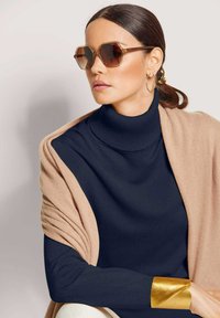 Frau mit dunklem Haar in einem tiefen Pferdeschwanz, trägt hexagonale braune Sonnenbrille, goldene Creolen, einen marineblauen Rollkragenpullover, einen beigen Schal und ein goldenes Armreif.