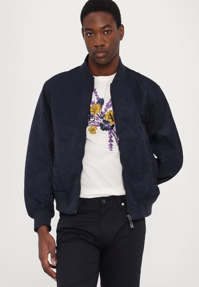 Navy bomber jacket with a shiny texture, zip closure, and ribbed cuffs, worn over a white t-shirt featuring floral embroidery in yellow and purple.