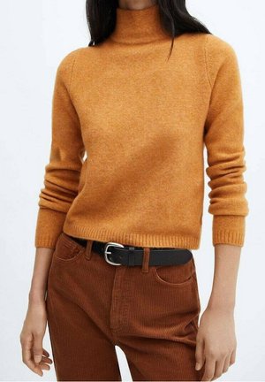 Kobieta w żółtym golfie o długich rękawach oraz wysokich brązowych sztruksowych spodniach z paskiem na czarnej klamrze.