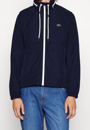 Marineblaue Zip-Up-Jacke mit weißen Kordeln und kleinem Krokodil-Logo auf der Brust, getragen mit blauen Jeans.