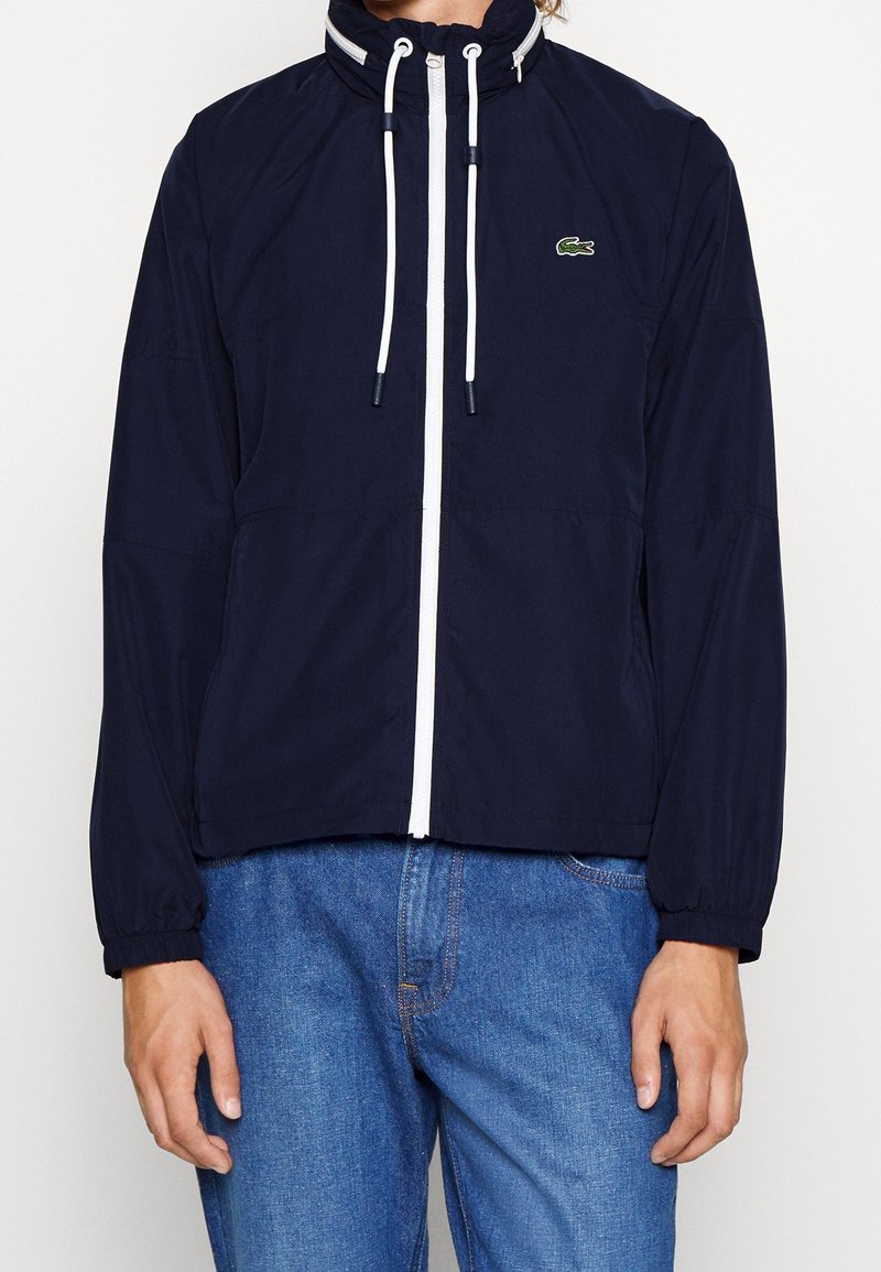 Veste zippée bleu marine avec des cordons blancs et un petit logo crocodile sur la poitrine, portée avec un jean bleu.