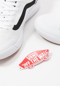 White sneakers with black accents, textured fabric, and a rubber sole. Includes a red Vans "Off the Wall" tag with white lettering.