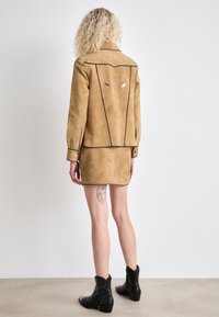 Giacca in suede beige con design strutturato, dettagli in borchie lungo l'orlo e i polsini, e un'unica decorazione sul retro. Abbinata a una gonna coordinata.