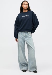 Ναυτικό φούτερ με κέντημα "Tommy Hilfiger", χαλαρή εφαρμογή και φαρδιά μανίκια; συνδυασμένο με ανοιχτό μπλε φαρδιά παντελόνια και μαύρα παπούτσια.