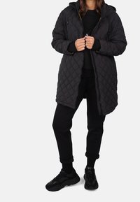 Černá prošívaná dlouhá bunda s kapucí, předním zipem a bočními kapsami, doplněná černými tepláky a teniskami. Hladká textura látky.