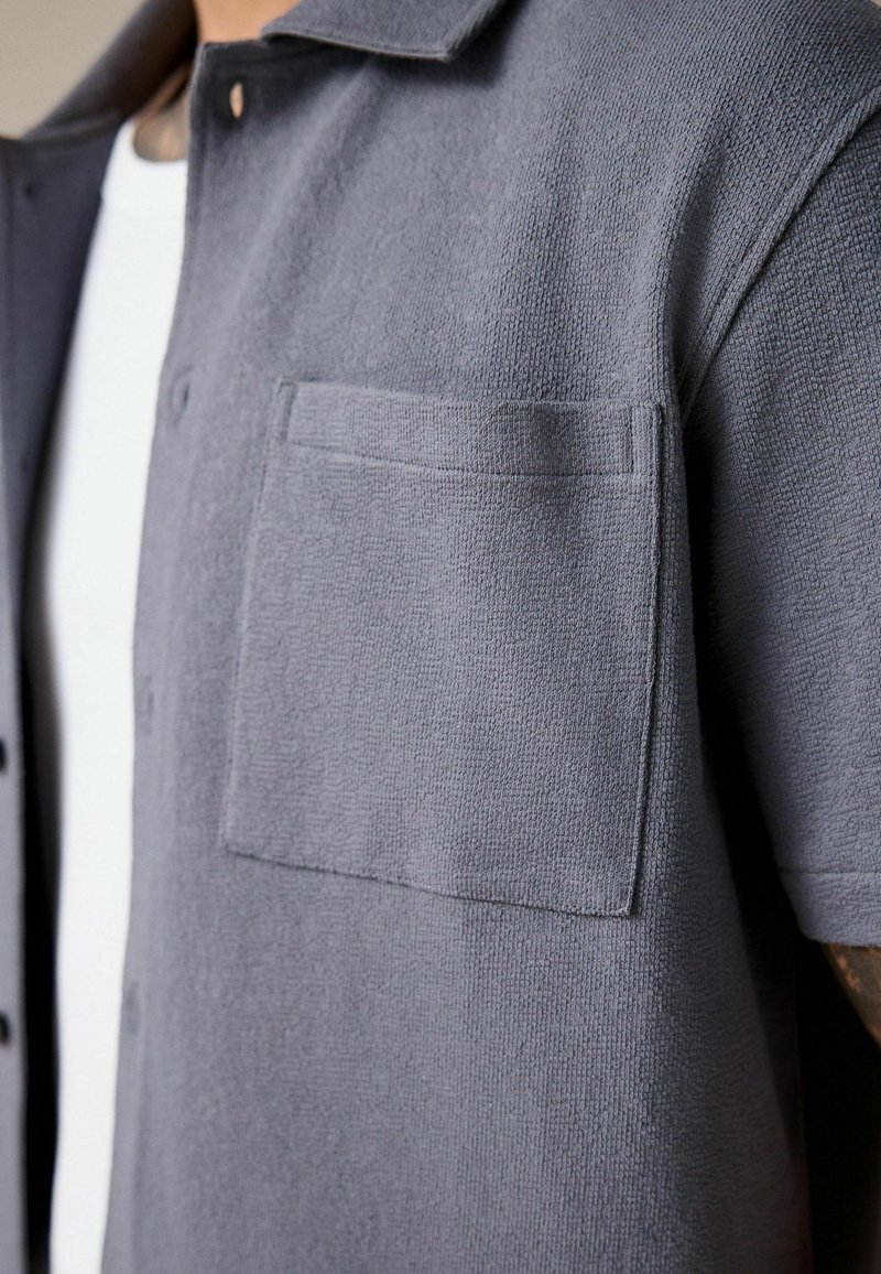Gros plan d'une personne portant une chemise grise texturée à manches courtes avec une poche poitrine par-dessus un t-shirt blanc.