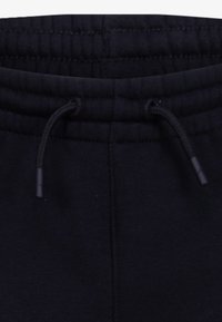 Černé teplákové kalhoty s elastickým pasem a šňůrkami s plastovými konci, švy jsou na tkanině viditelné.