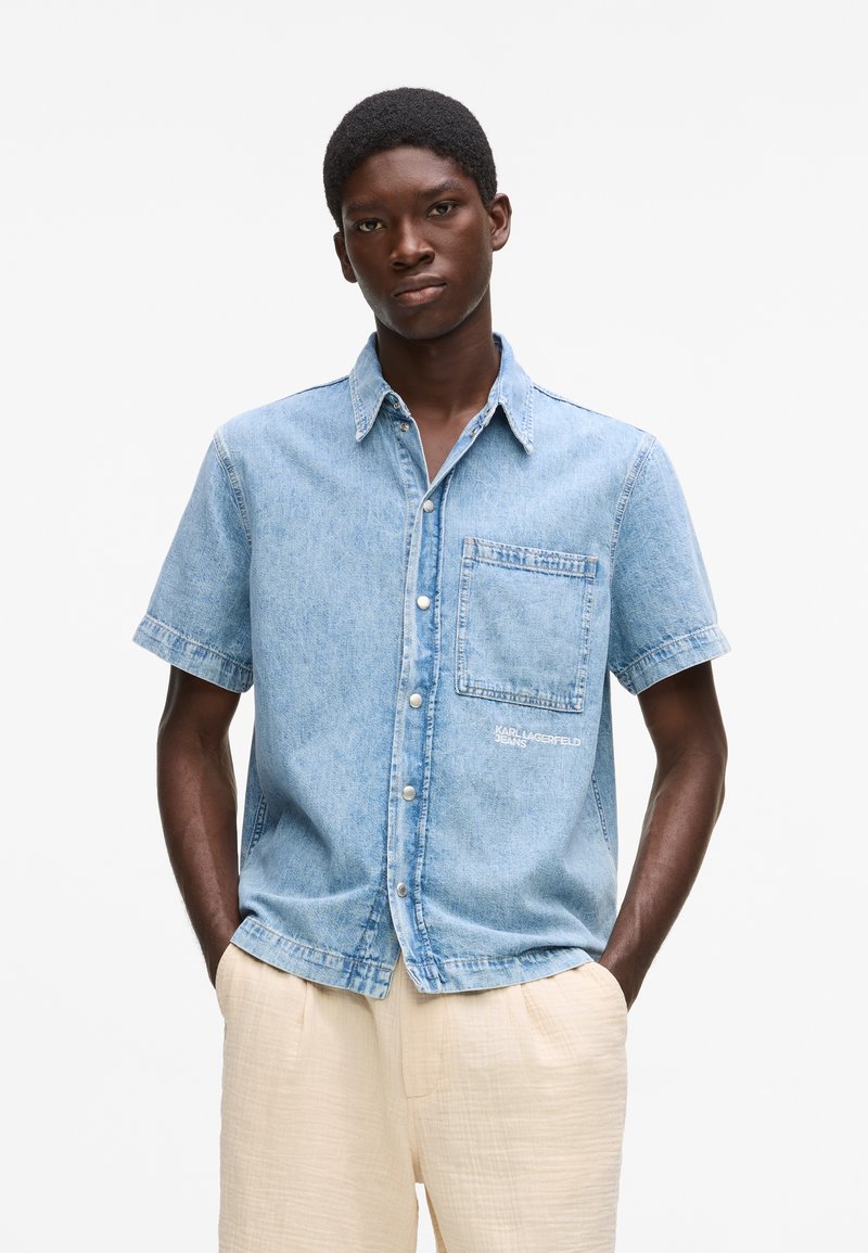 Νεαρός άνδρας που φοράει ένα ανοιχτό μπλε κοντομάνικο τζιν πουκάμισο με τσέπη στο στήθος και μπεζ παντελόνι, στέκεται με τα χέρια στις τσέπες.