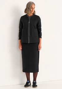 Kvinna med axellångt grått hår iförd en svart zip-up jacka, svart medellång kjol, svarta strumpbyxor och svarta sneakers med vita ränder.