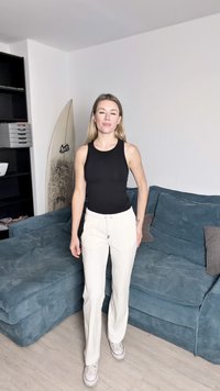 Melns ribots topiņš, balti bikses ar auklu un balti sporta apavi. Fonā zils teksturēts dīvāns, pie kura atbalstīta sērfošanas dēlis.