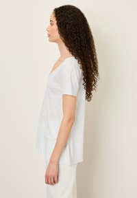 Bijela majica kratkih rukava s dubokim V-izrezom, lepršavom kroju i blago zakrivljenim rubom. Lagani, mekani materijal. Model stoji u profilu.