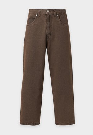 Jeune homme debout portant une chemise bleue boutonnée, un pantalon marron à jambes larges et des baskets blanches sur un fond clair uni.