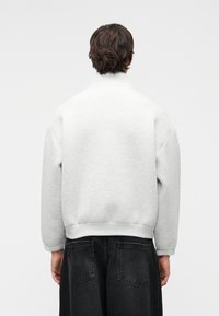 Άτομο με κοντά καστανά μαλλιά που φοράει γκρι ανοιχτό sweatshirt με ψηλό γιακά και φαρδιά μαύρα τζιν, από πίσω με φόντο έναν απλό τοίχο.