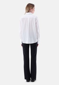 Camicia bianca a maniche lunghe con colletto testurizzato, abbinata a pantaloni neri svasati. Tessuto morbido, taglio classico e vestibilità sartoriale.