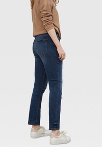 Mujer vestida con pantalones ajustados de mezclilla azul oscuro, suéter beige y zapatillas blancas, de pie de lado sobre un fondo claro y liso.