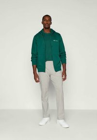 Haut zippé vert avec cordons et logo, associé à un pantalon tapered gris clair. Le mannequin porte des baskets blanches, se tenant sur un fond neutre.