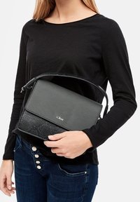 Vrouw in een zwart shirt met lange mouwen en blauwe jeans, die een zwarte s.Oliver handtas vasthoudt met een getextureerde klep en schouderriem.