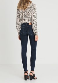 Mujer vestida con jeans skinny de color azul oscuro, blusa de estampado de leopardo y sandalias de tacón negro, de pie de espaldas sobre un fondo liso.