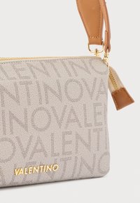 Bež Valentino torbica za zglob s monogram uzorkom, zlatnim patentnim zatvaračem i tamnobraon kožnom naramenicom i povlačnim jezičkom patentnog zatvarača.