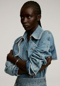 Chaqueta de denim en azul claro con un collar estructurado, mangas holgadas y detalles de botones; cuenta con una cintura ceñida y bordes deshilachados.