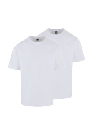 Jeune homme aux cheveux courts portant un t-shirt blanc uni et un jean bleu clair, debout devant un fond gris neutre.