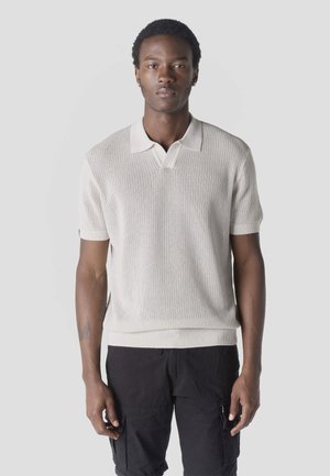 Fiatal férfi rövid, göndör hajjal, krémszínű kötött rövid ujjú pólót és fekete cargo rövidnadrágot visel, világosszürke háttér előtt áll.