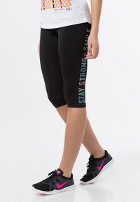 Leggings capri neri con il testo "STAY STRONG" in lettere colorate e riflettenti lungo il lato. Indossati con scarpe da ginnastica nere e rosa.