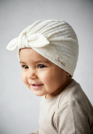 Smiling baby wearing a textured white turban-style hat with a bow and beige clothing against a plain light background.