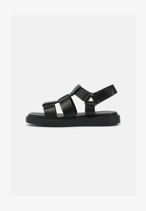 Sandalia de cuero negro con múltiples correas anchas, suela plana y correa ajustable en el tobillo. Textura suave y diseño minimalista.