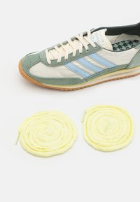 Sportsko sko med grønn semsket skinn og krem tekstiloverdel, blå striper og såle i gummigummi. To gule, fluffy runde lisser inkludert.