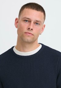 Man with short brown hair and blue eyes wearing a navy blue knitted sweater over a white shirt, facing forward with a neutral expression.