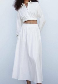 Blouse blanche courte à boutons et manches longues, assortie à une jupe midi blanche fluide avec une taille élastique et un léger éclat.