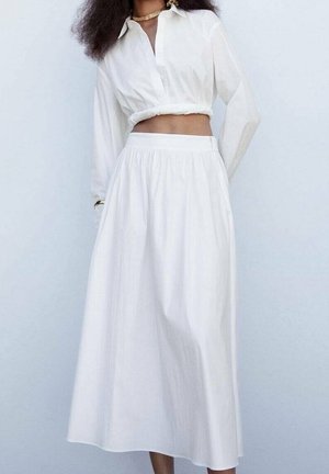 Blouse blanche courte à boutons et manches longues, assortie à une jupe midi blanche fluide avec une taille élastique et un léger éclat.